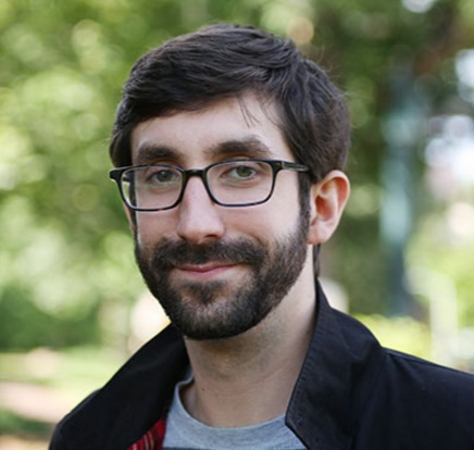 Stephen Capuzzi is a young man with dark hair, a beard, and glasses. He is standing in front of blurred trees with a dark jacket.