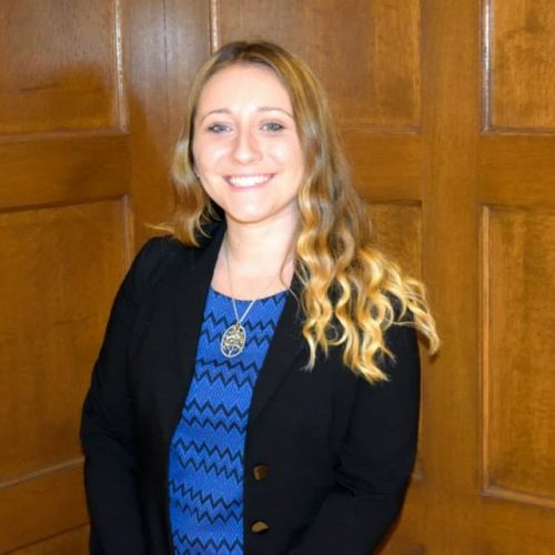 Ashley Crandall is smiling in front of wooden panels. She has blonde hair. She is wearing a dark cardigan with a blue, patterned top, and a gold necklace.