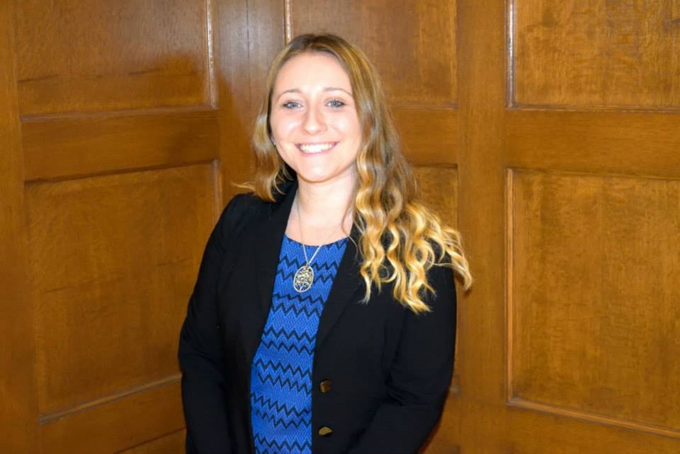 Ashley Crandall is smiling in front of wooden panels. She has blonde hair. She is wearing a dark cardigan with a blue, patterned top, and a gold necklace.