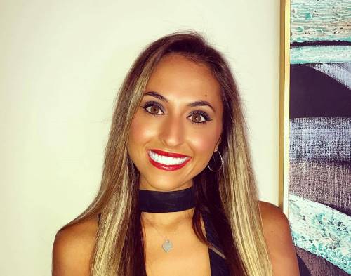 Rachel Brida is smiling in front of a wall with a print. She is wearing red lipstick and a black top with a silver necklace.