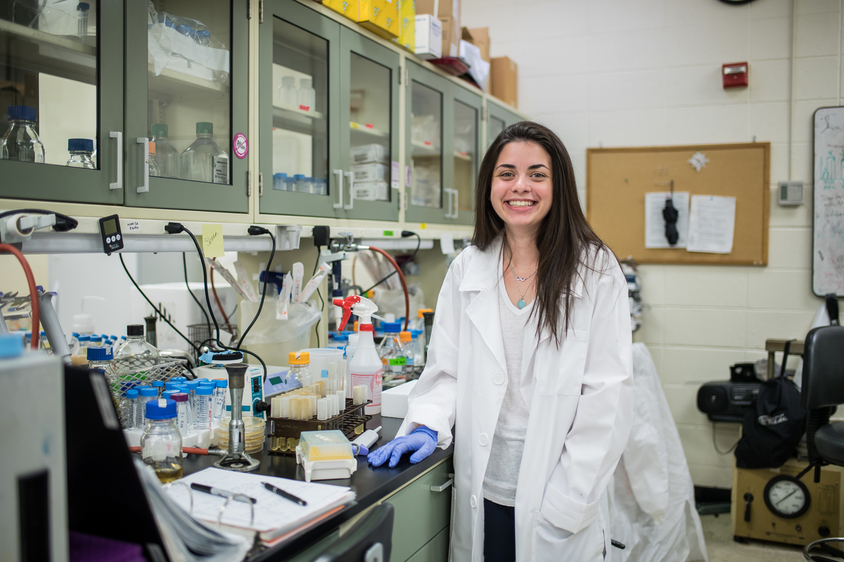 Marisa Egan is smiling while standing next to a counter in a lab. She is wearing a white lab coat and rubber gloves.