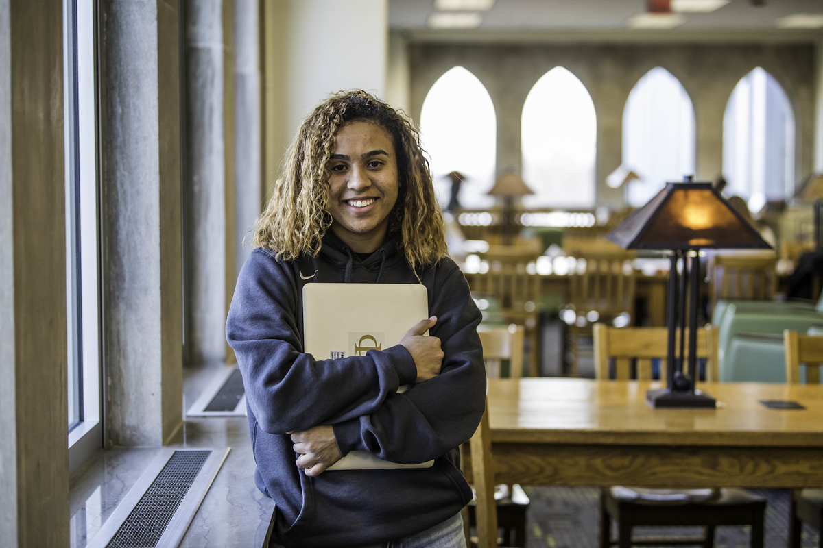 Vilma Fermin smiles through her curly hair and wears a dark grey SJU sweatshirt. She holds her silver computer to her chest in the library.