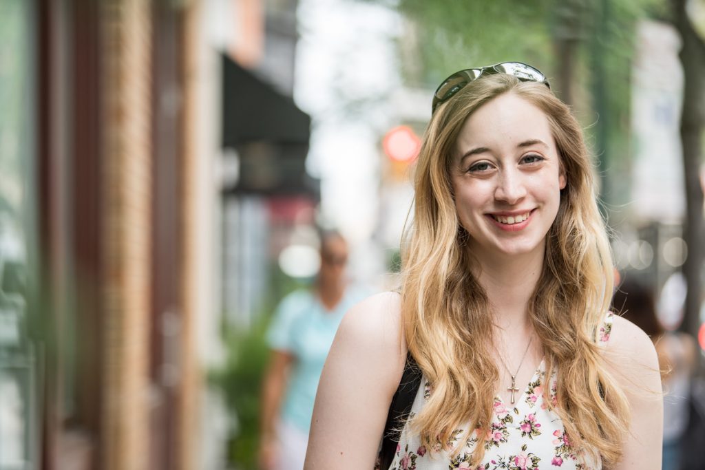Elana Valentin smiles on a busy Philadelphia street. She wears a white shirt with flowers, silver sunglasses on her head and a black backpack.