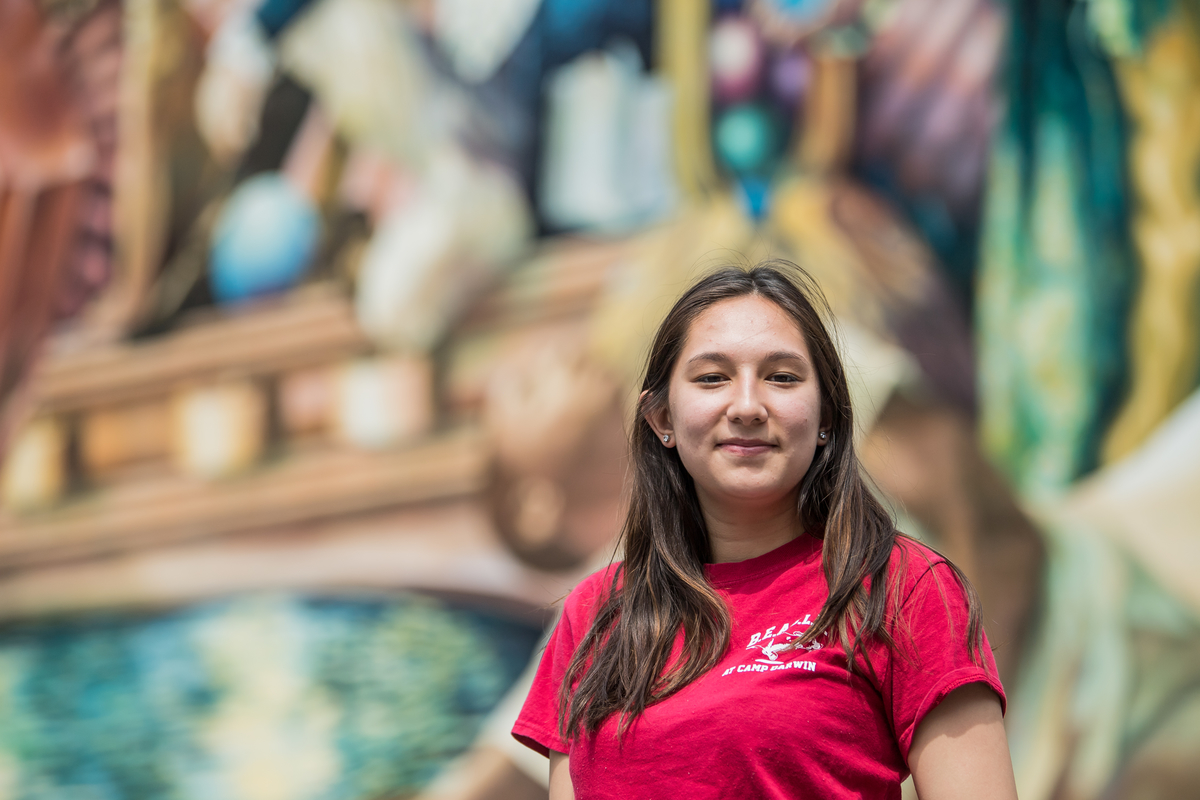 Ave Burleigh wears a red t-shirt with the SJU logo and smiles in front of a colorful mural.