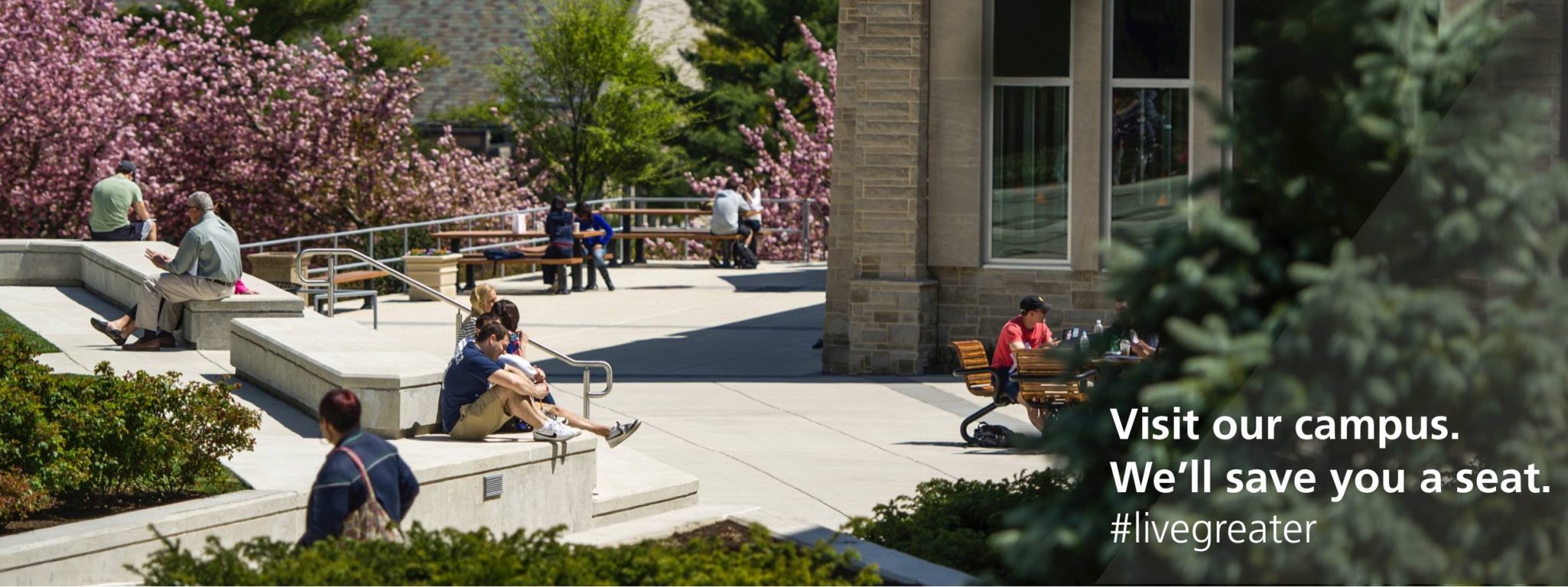 Students sit in the sunshine through the SJU quad. The screen reads, "Visit our campus. We'll save you a seat. #livegreater"