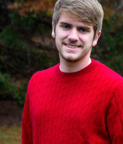 Charles Gallagher wears a red sweater and smiles before a canopy of fall leaves.