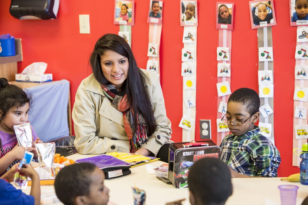 Gabriela sits in a classroom with young students, a red wall behind her