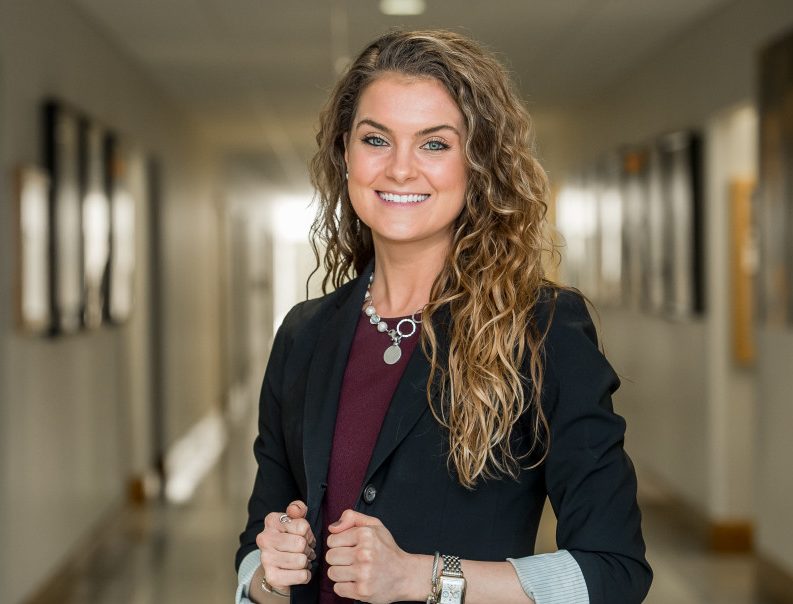 Ashley Wolf is smiling in a blurred hallway. She has curly, long hair, and is wearing a dark blazer, dark red top, and a white necklace.